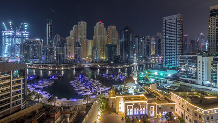 Obraz premium Yachts in Dubai Marina flanked by the Al Rahim Mosque and residential towers and skyscrapers aerial night timelapse.