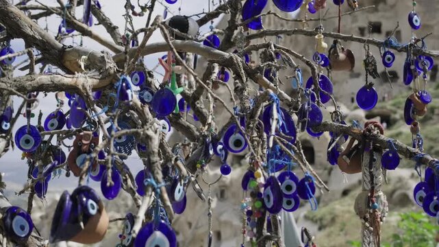 Several nazar boncuk, traditional blue turkish evil eye charms, are suspended from the tree branches as protective talismans, set against the picturesque backdrop of the rocky cappadocia landscape