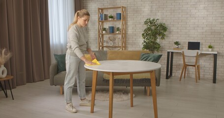 A woman in casual attire is cleaning a round table in a stylish living room with a cozy atmosphere.