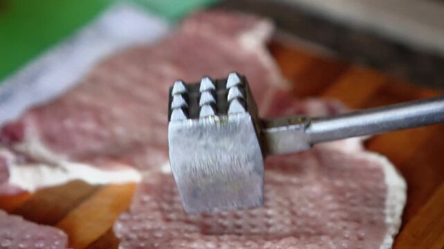 A woman tenderizes fresh raw pork chops with a metal mallet on a wooden cutting board. The process of cooking a homemade meal in the kitchen. The process of cooking schnitzel. 