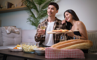 Smiling couple sitting on a sofa enjoying pizza and wine at home.