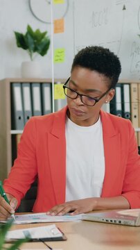 Afro american woman office manager which sitting in front of computer and previsioning report and noticing needed details into papers