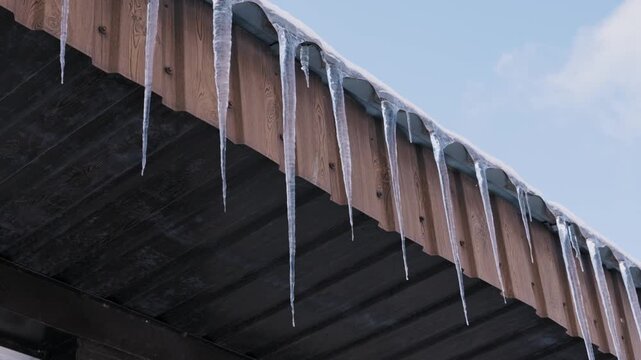 Long, sharp icicles hang in a row from the edge of a wooden roof. The low-angle shot captures the frozen water against a clear blue winter sky.