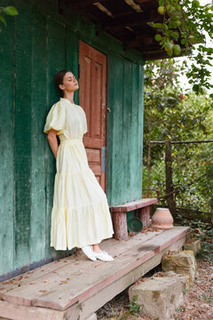 Woman in a pale yellow dress leaning against a green wooden wall outdoors, serene expression, sunlit garden setting creating a calm, timeless mood with rustic doorway and casual elegance