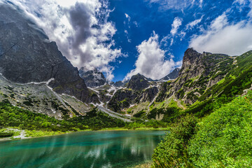 A stunning wide panorama of Zelene pleso (Green Lake) with the historic mountain hut and jagged granite peaks reflecting in the crystal clear turquoise water. High Tatras National Park, Slovakia.