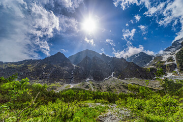 Obraz premium A powerful mountain landscape featuring high granite walls with snow patches under a bright sun with rays. Lush green bushes and rocky terrain in the foreground, located in the Tatra National Park, Sl