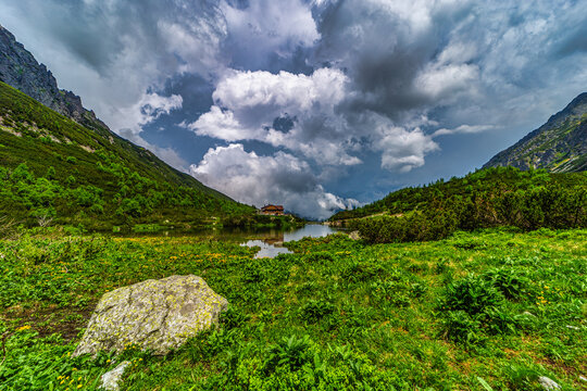 A scenic view of the mountain cabin "Chata pri Zelenom plese" nestled in a lush green valley. Surrounded by high rocky peaks and dark, moody storm clouds in the High Tatras National Park, Slovakia.