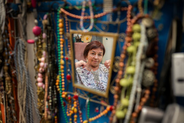 A woman looks at her reflection in a small mirror framed by colorful necklaces, trying on jewelry in a market stall, surrounded by textures, colors, and handmade details.