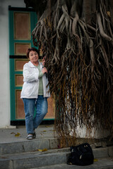 A woman stands beside massive hanging roots of an old tree near a historic building, blending human presence with organic forms in a quiet urban courtyard.