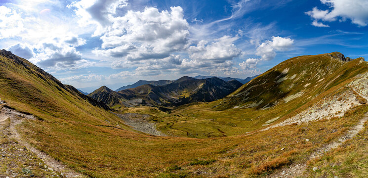 A narrow rocky path winds up a steep grassy slope toward a mountain summit. Scenic view from Hladke sedlo in High Tatras National Park, featuring a turquoise lake in the valley under a cloudy blue sky