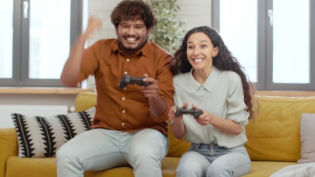 A couple is sitting on a yellow sofa, holding game controllers and smiling widely. They are engaged in a gaming session in a bright and inviting living room with plants.