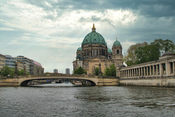 Fototapeta premium Berlin Cathedral and Spree River Boat Tour - A tourist boat cruises on the Spree River past the iconic Berlin Cathedral (Berliner Dom) under a dramatic, cloudy sky with beautiful sun rays.