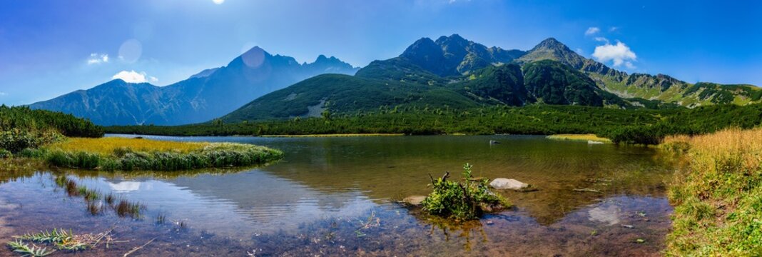 A stunning wide panorama of the crystal clear Velke Biele pleso mountain lake. Surrounded by majestic peaks like Jastrabia veža and lush green slopes under a clear blue sky in the Tatra National Park.