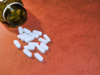 White Pills Spilled from Bottle - White oblong pills spilled from an empty amber medicine bottle on a textured red-orange background, symbolizing health and treatment.