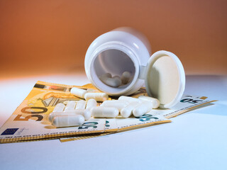 High Cost of Medicine in Euros - Overturned white pill bottle with capsules scattered on multiple 50 Euro banknotes, highlighting the financial burden of medication and healthcare costs.