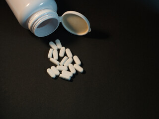 Pills Spilled from Open Medicine Bottle - A scattering of white oval pills, many marked IH25, have fallen from an open white plastic medicine bottle onto a dramatic dark surface. Medical concept.