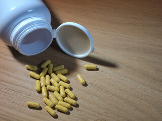 Medicine Pills Spilled on Wood Table - Yellow capsule pills scattered from an overturned white prescription bottle on a light brown wooden surface. Health, pharmacy, and medication concept.
