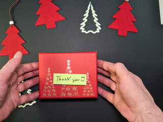 Thank You Gift Box with Christmas Decor - Overhead view of hands presenting a red holiday gift box adorned with golden Christmas trees and a handwritten 'Thank You' message on a dark background.