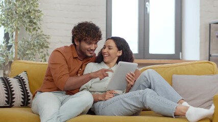 A cheerful couple sits comfortably on a yellow couch in a cozy living room. They smile and laugh as they look at a tablet together, sharing a joyful moment in their home.