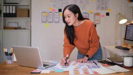 Focused woman standing at her desk designing wireframes for a mobile app interface using printed UI elements.