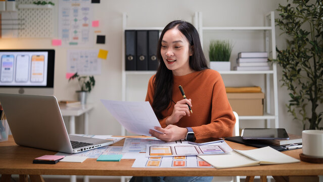Female UX designer presenting wireframe sketches for a mobile app while sitting at a desk in a creative workspace. - Powered by Adobe