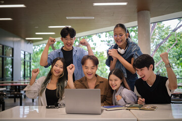 Obraz premium Group of excited university students cheering together around a laptop after achieving success on a project or exam result in a modern campus study area.