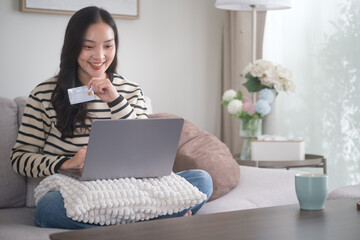 Smiling woman shopping online from home, holding a credit card while using a laptop on a sofa. Online payment and e-commerce lifestyle concept.