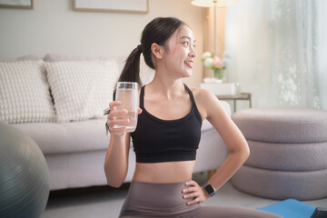 Young woman holding a glass of water, relaxing after exercise on a yoga mat. Healthy lifestyle, home fitness, and wellness concept