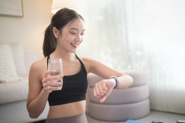 Smiling woman checking her fitness tracker while holding a glass of water at home. Home workout, hydration, and healthy lifestyle concept.