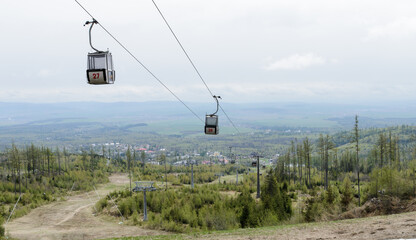 Cable car gondolas numbered 27 and 38 ascending mountain slope in Tatranská Lomnica, Slovakia. Scenic alpine landscape with forest, ski runs, and valley town view under cloudy sky