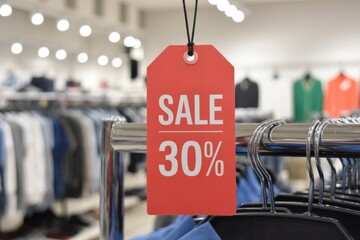 Red sale tag with 30 percent discount hanging on clothing rack in store