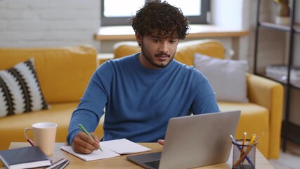 A man is seated at a wooden desk in a cozy living room, writing in a notebook. He has a laptop open and a cup of coffee nearby, focusing on his task with a thoughtful expression.