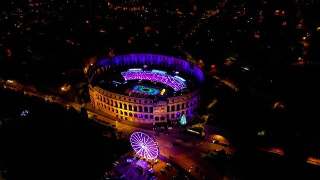 Aerial view of the ancient Pula Arena illuminated with vibrant lights and a festive Ferris wheel during Advent in Pula, Croatia. A magical winter night scene.