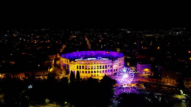 Aerial view of the ancient Pula Arena illuminated with vibrant lights and a festive Ferris wheel during Advent in Pula, Croatia. A magical winter night scene.