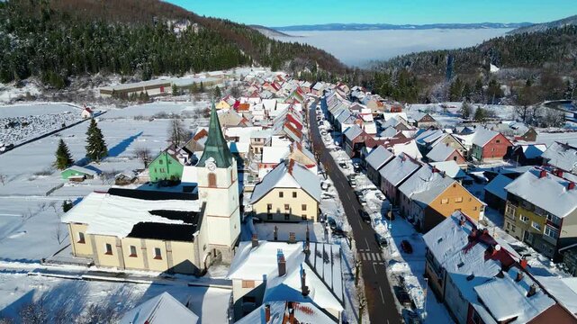 Winter's embrace in a charming Croatian village Delnice, with snow-dusted roofs and a prominent church under a clear blue sky. Fog blankets the distant valley.