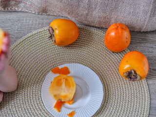 Fresh Ripe Persimmons on a Woven Placemat with a Sliced Fruit on a White Plate - Overhead View of Vibrant Orange Persimmons and Fruit Slices for a Healthy Snack