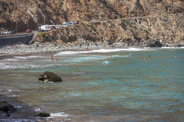 Busy beach scene with swimmers, parked cars along coastal road, and rugged volcanic cliffs at Roque de las Bodegas, Tenerife, Canary Islands, Spain.