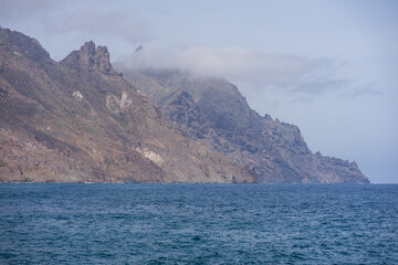 Misty volcanic mountain peaks and rugged cliffs rise above deep blue ocean horizon at Roque de las Bodegas, Tenerife, Canary Islands, Spain.