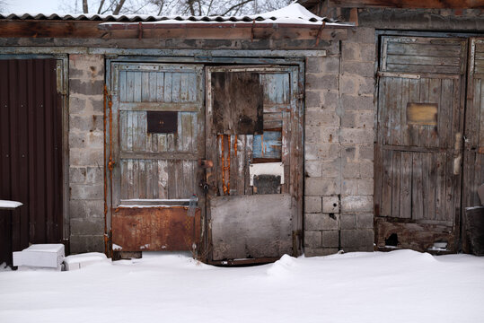 Weathered wooden garage doors in winter. Old abandoned shed with peeling paint and rusty metal patches covered in snow.