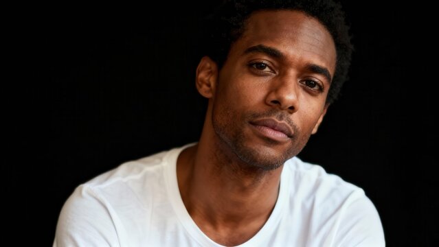 Serious young adult Black man wearing a white t-shirt captured in a dramatic high contrast  portrait against a black background.