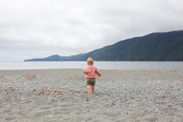 A toddler plays along a sandy beach in the on a cloudy day. Beach days provide important unstructured play opportunities for children to engage with their environment.