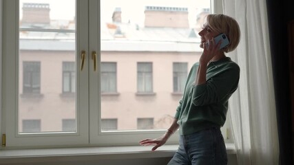 Positive happy middle aged woman talking on smartphone with friend, smiling and joking, enjoying friendly conversation and sharing gossips. Female chatting on phone, standing beside window in room.