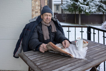 Senior male sitting outside house reading print media during breakfast in warm clothes. Quiet moment on lifestyle scene.