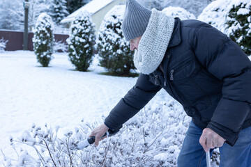 Elderly man in warm clothing trimming garden shrubs with pruners using cane for support in snowy outdoor yard.
