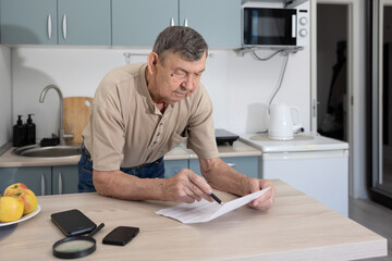 Senior male examining household payment document at home kitchen with smartphone and magnifier on table.