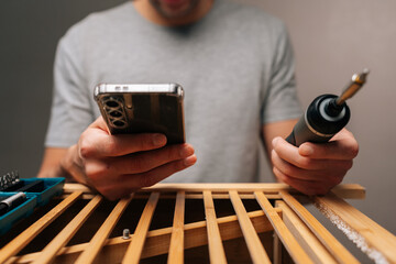 Cropped shot of handyman assembling wooden furniture, holding cordless electric screwdriver and checking instructions on smartphone for diy home improvement and renovation project.