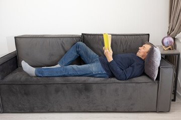Elderly man lying on sofa reading book in calm living room environment emphasizing leisure and quiet indoor lifestyle.