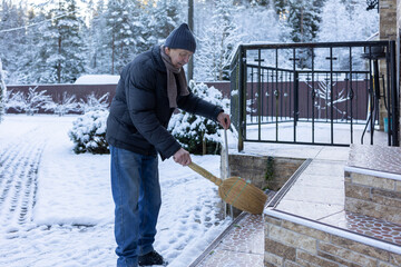 Adult man in winter clothing sweeping snow from porch steps with broom, outdoor home area in cold season maintenance task.