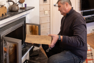 Senior male loading wooden log into stove, lights fireplace inside living. Space winter warmth concept.
