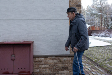 Senior male moving carefully with walking stick on brick pavement surrounded by snow covered landscape cold season daily activity.